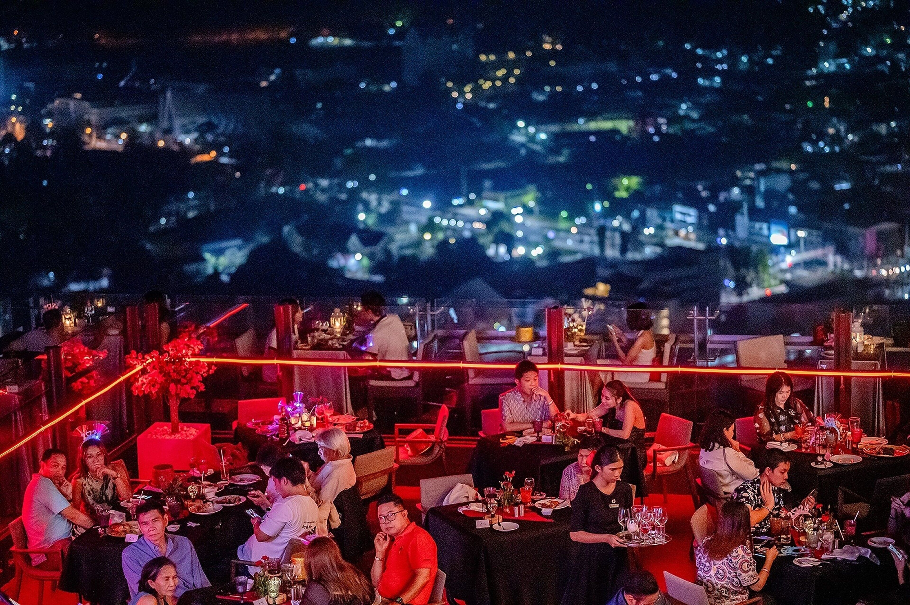 Un restaurant avec des tables dressées pour le dîner, une vue sur le paysage urbain illuminé la nuit et un arbre décoratif au premier plan.