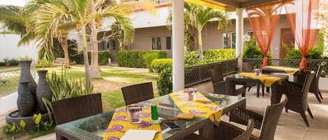 An outdoor dining area with tables and chairs, surrounded by greenery and palm trees.