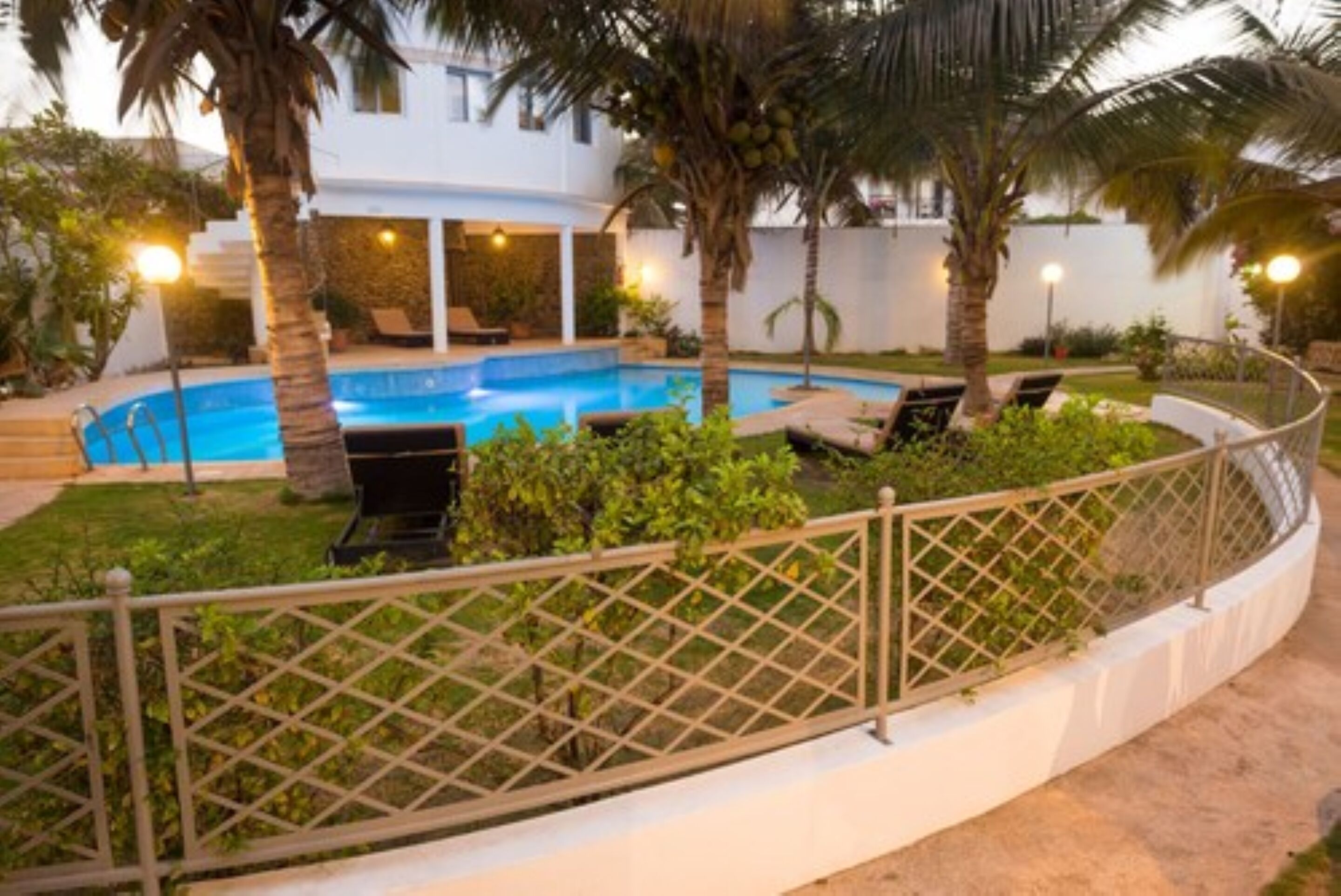 A swimming pool surrounded by palm trees and lounge chairs, with a white building in the background.