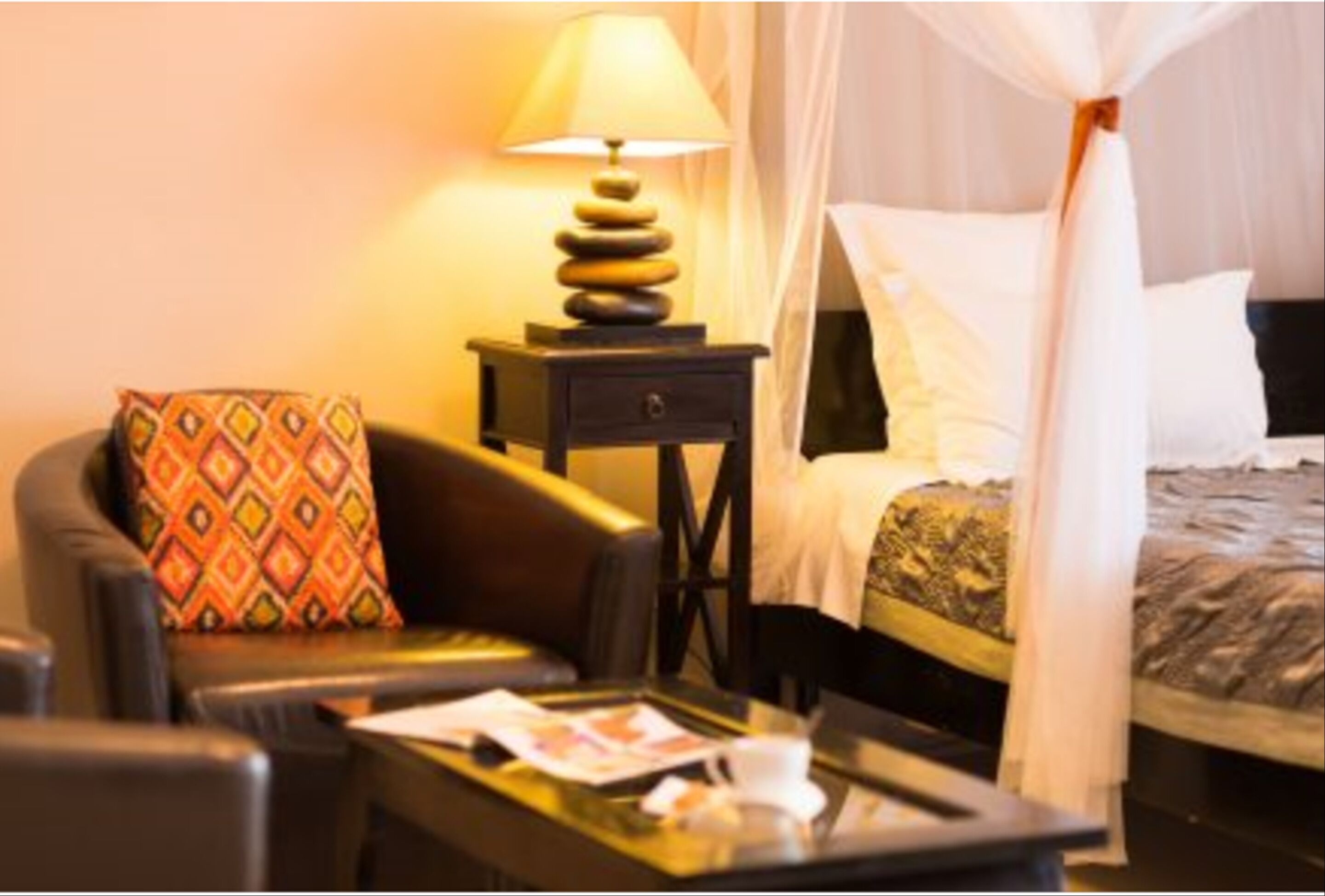 A hotel room with a bed, a bedside table with a lamp, a sofa with a patterned cushion, and a glass coffee table.