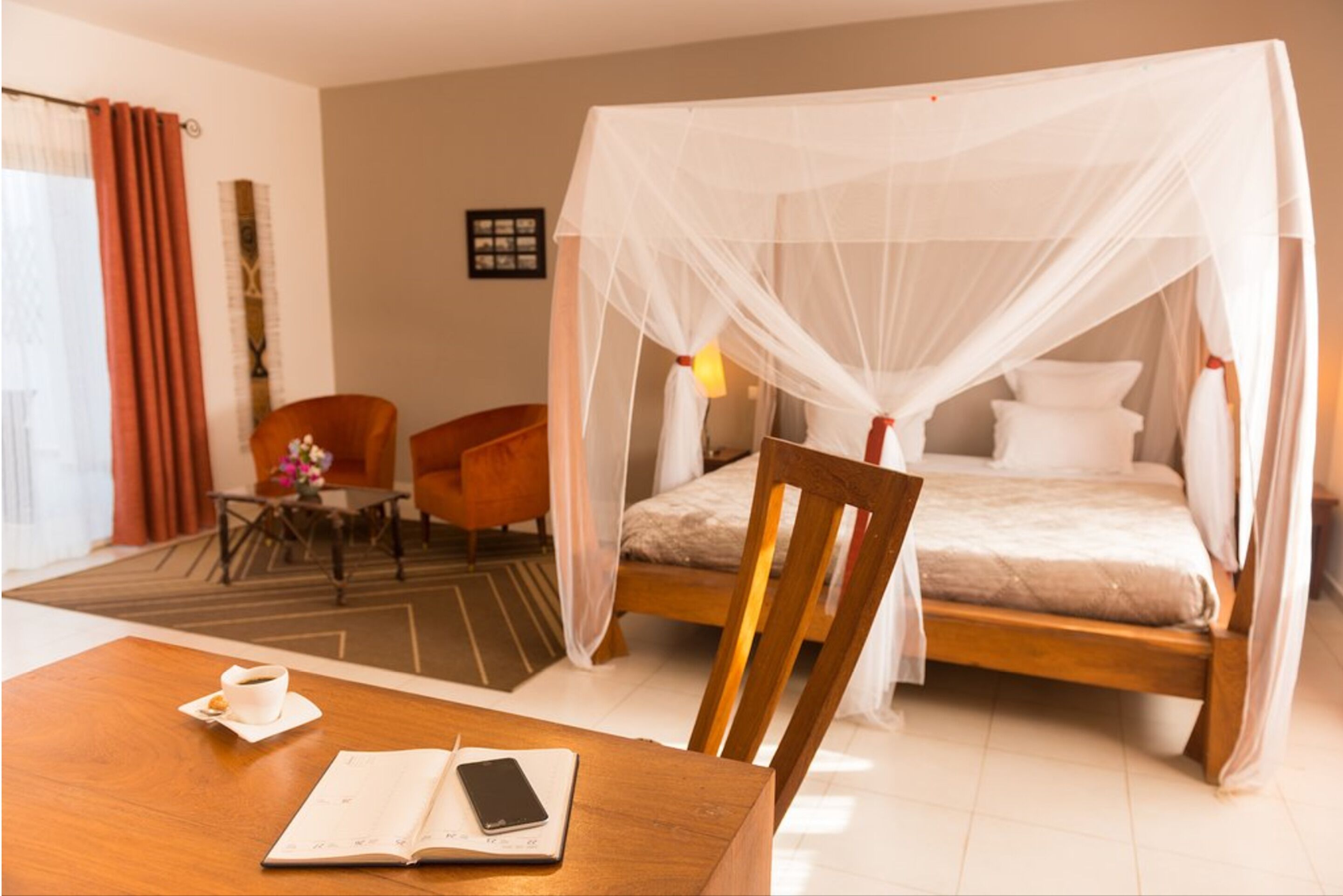 A hotel room with a wooden bed, a wooden chair, a small table with a cup and a book, and a mosquito net over the bed.