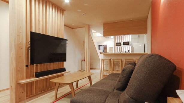 Een moderne woonkamer met een flatscreen televisie, houten paneelwanden, een bruine bank en een keuken op de achtergrond.