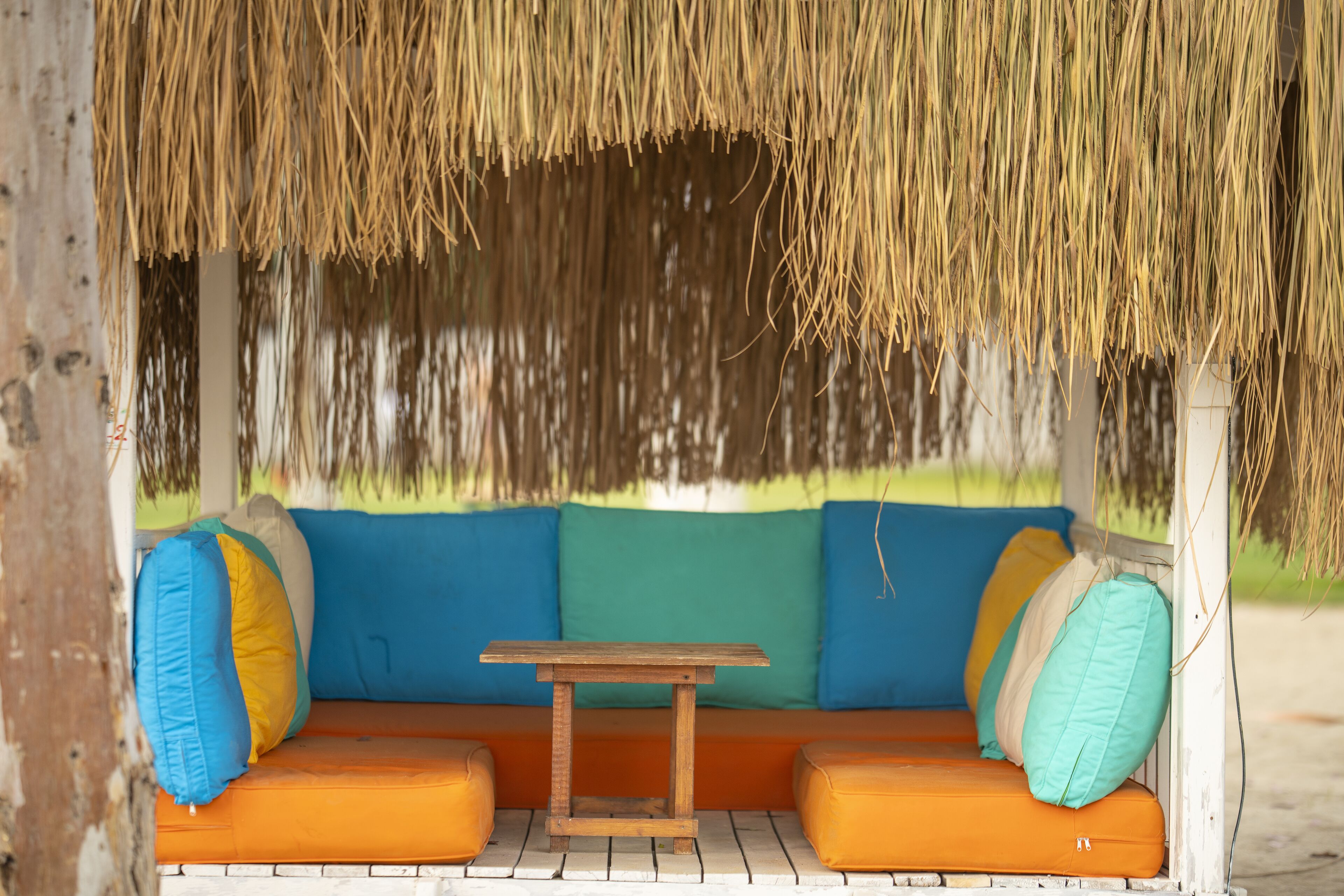 Estructura de techo de paja con una mesa de madera y asientos acolchados color naranja, decorados con cojines de colores.