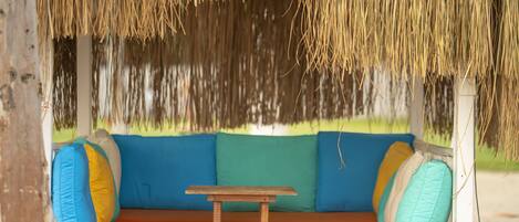 Estructura de techo de paja con una mesa de madera y asientos acolchados color naranja, decorados con cojines de colores.