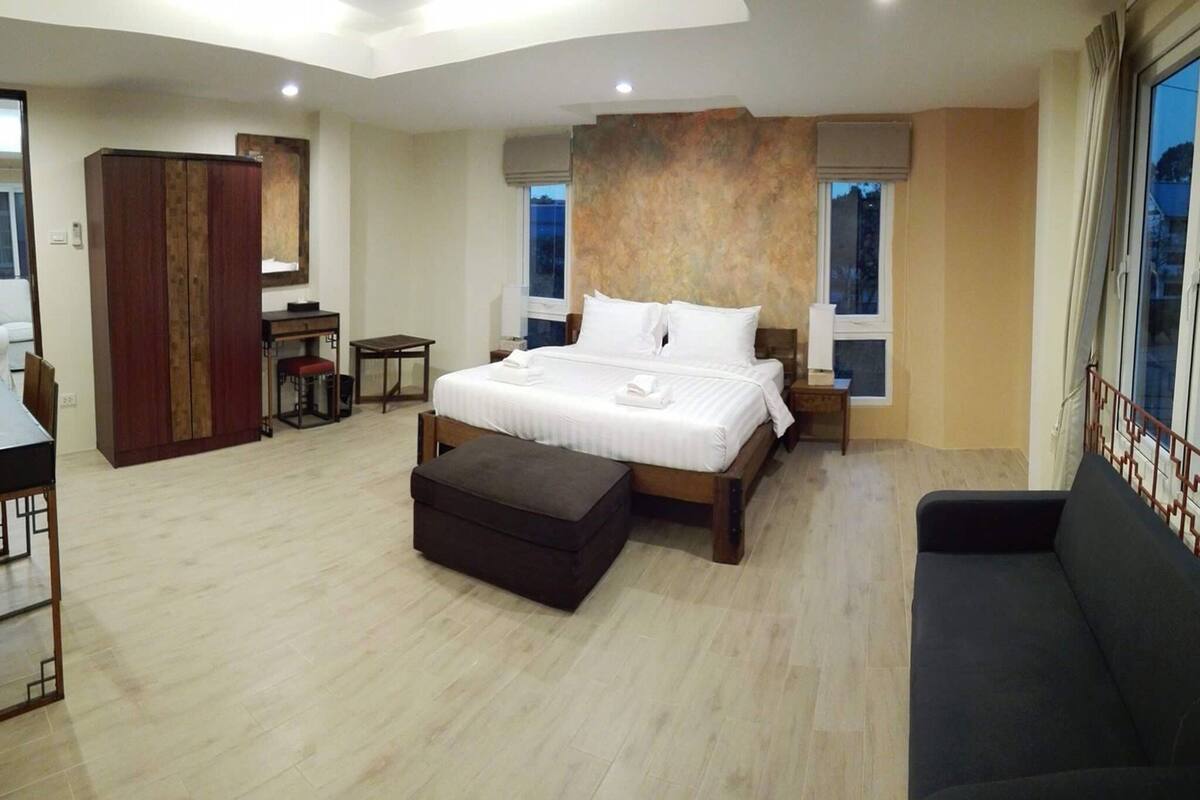 A spacious bedroom with a large bed, a dark sofa, a wooden wardrobe, a desk, and a small table.