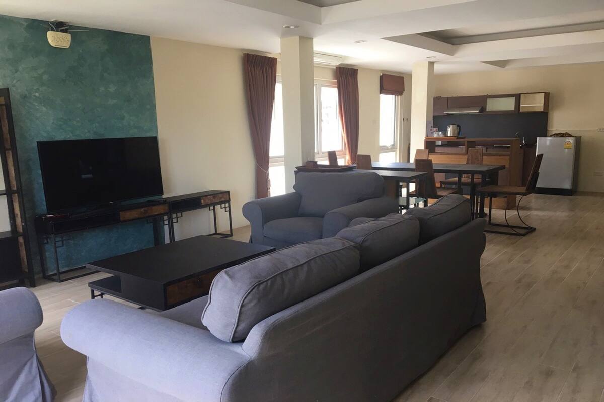 A modern living room with a large grey sofa, a flat-screen TV, and a dining area with a table and chairs.