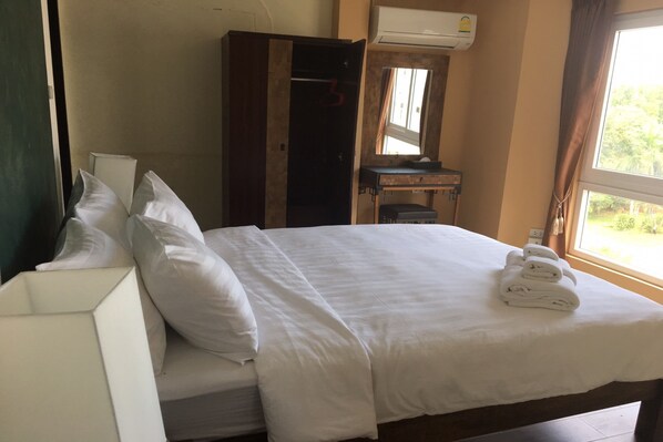 Una cama bien hecha con ropa blanca y toallas, un armario de madera, un escritorio pequeño con espejo y una ventana con cortinas.