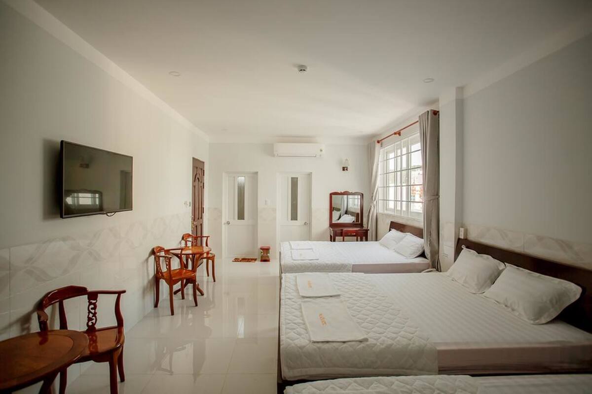 A hotel room with two beds, a TV mounted on the wall, a dining area with chairs, and a window with curtains.