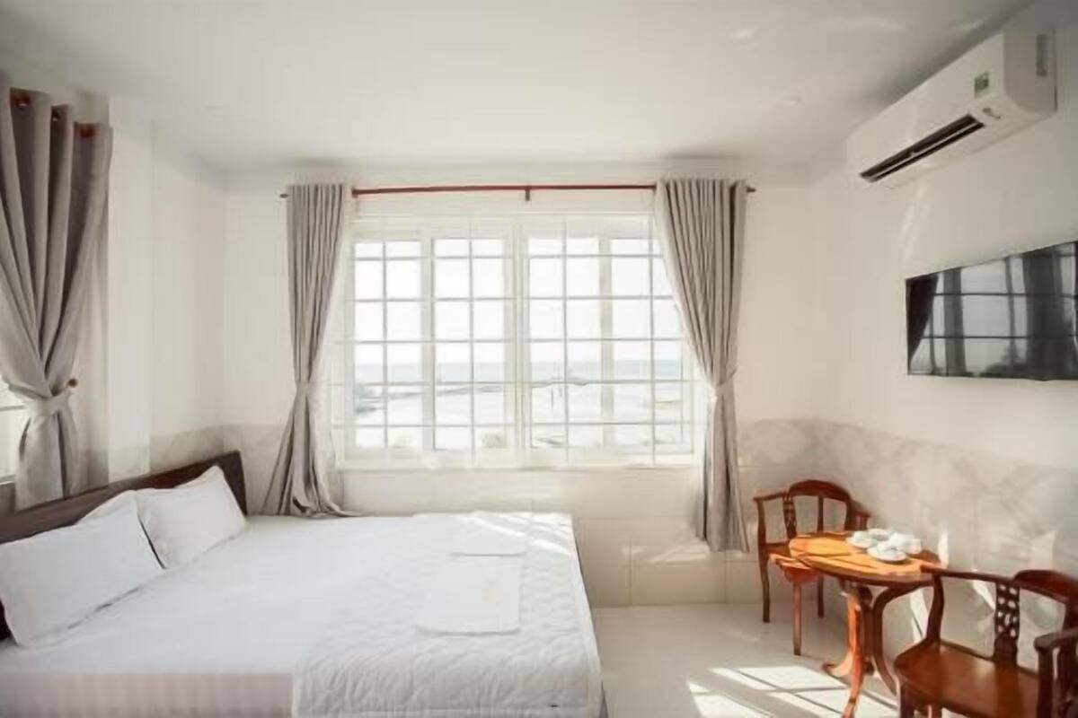 A hotel room with a bed, a dining table with chairs, a TV mounted on the wall, and a window with curtains.