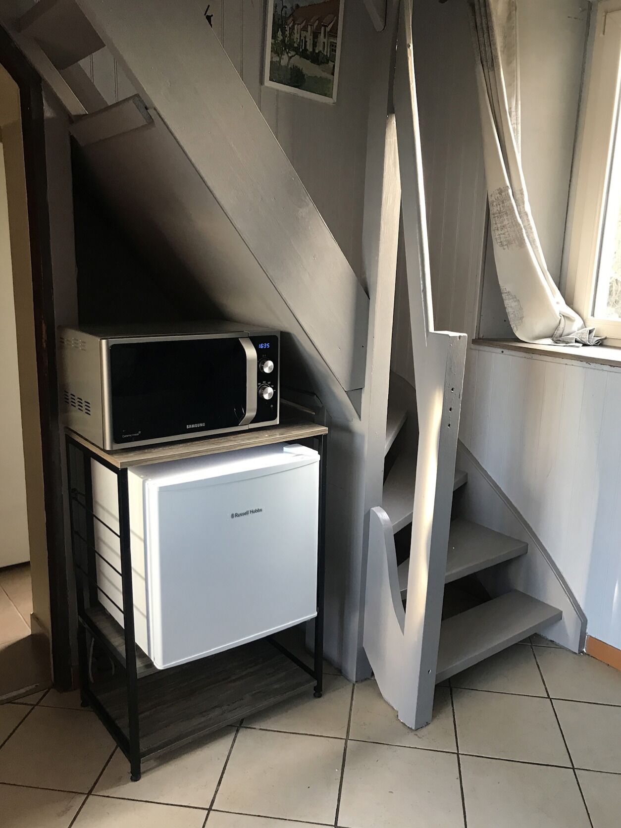 A compact kitchen area with a microwave on top of a refrigerator, a staircase leading to an upper level, and a window with curtains.