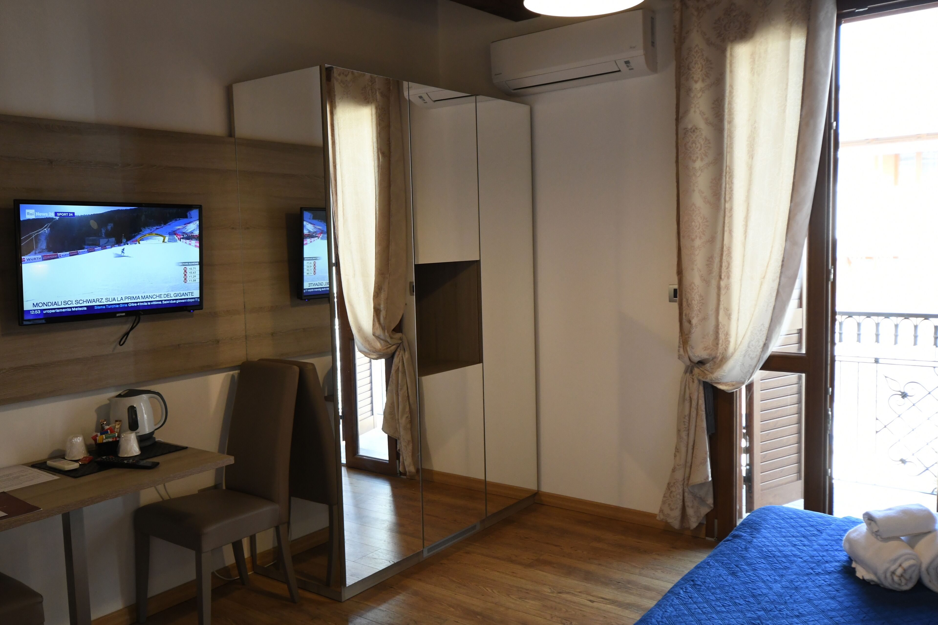 A hotel room with a TV on the wall, a desk with a kettle, a chair, a bed with towels, and a wardrobe.