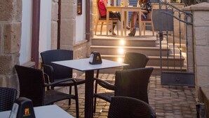An outdoor café with wicker chairs and tables, stone walls, and a staircase leading to a balcony with potted plants. - Double B - Maison De Charme (Alghero)