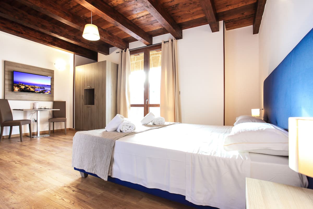 Habitación de hotel con techo de madera, cama con ropa blanca, escritorio con sillas y televisor en la pared.