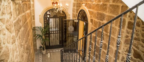 A stone-walled staircase with a wrought iron railing, leading to a door with a 'B' sign, and a chandelier above.