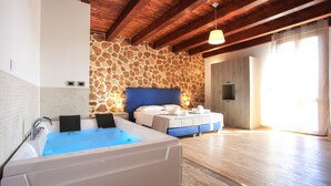 A spacious room with a large bathtub, a bed with a blue headboard, stone wall, wooden beams, and a large window. - Double B - Maison De Charme (Alghero)