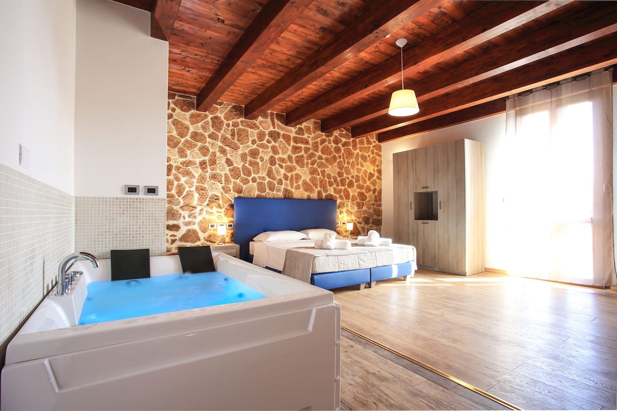 Una habitación amplia con una bañera grande, una cama con cabecera azul, pared de piedra, vigas de madera y un ventanal.