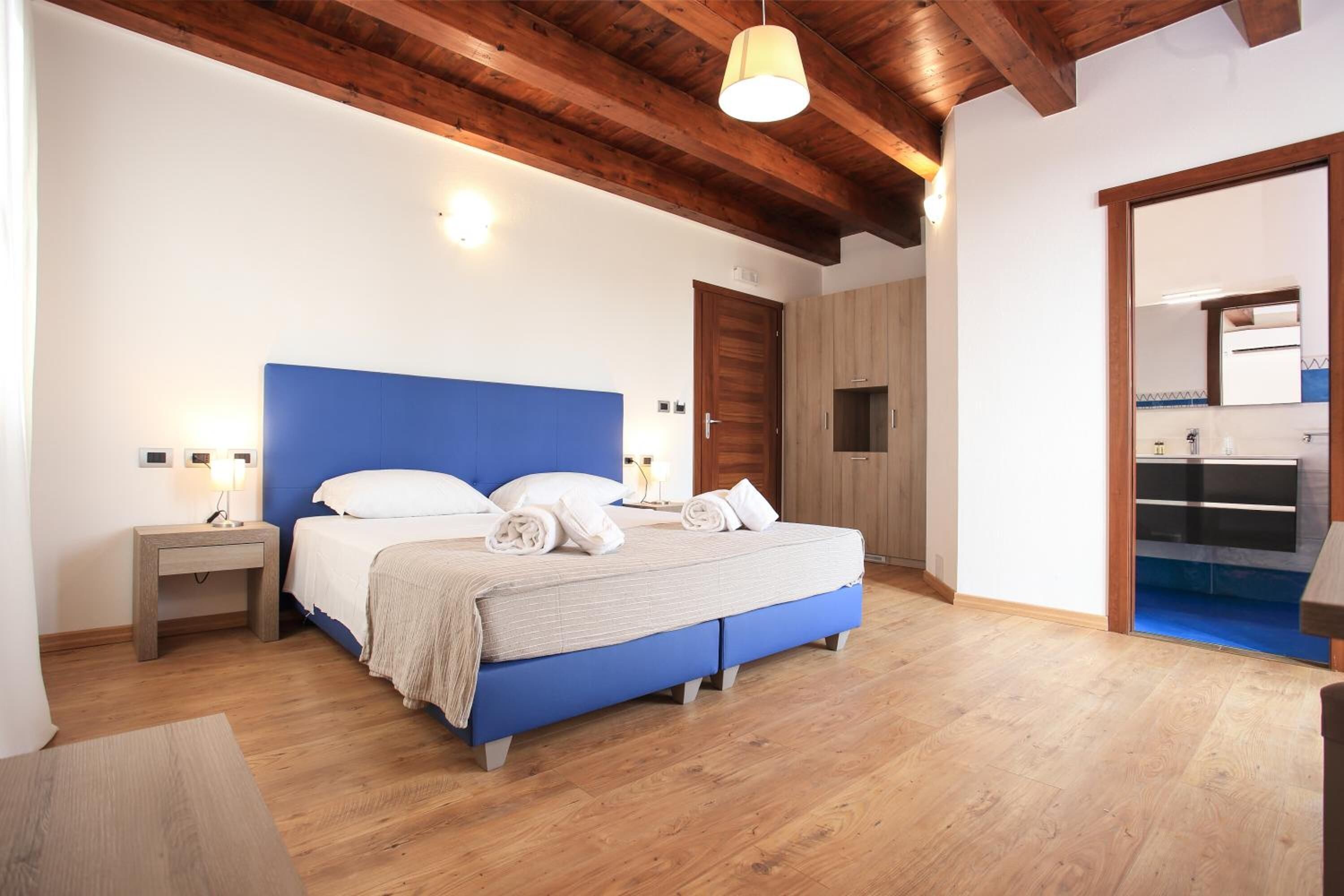 Ein Schlafzimmer mit blauem Kopfteil, Holzboden, einem Nachttisch mit Lampe und einem durchgehenden Türbogen einsehbarer Badezimmer.