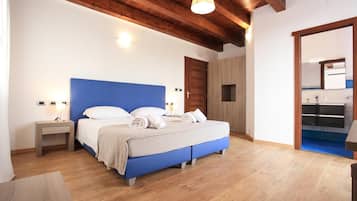Un dormitorio con cabecera azul, suelo de madera, mesita de noche con lámpara y baño visible a través de una puerta abierta.