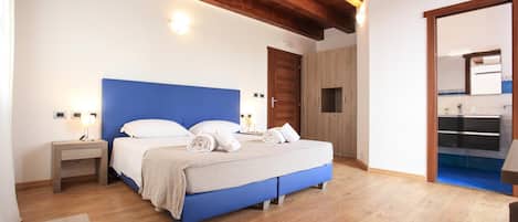 Un dormitorio con cabecera azul, suelo de madera, mesita de noche con lámpara y baño visible a través de una puerta abierta.