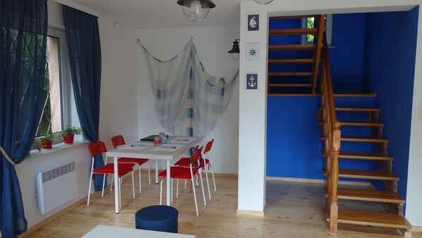 A room with a wooden staircase, a dining table with red chairs, a hammock, and a view of the outdoors.