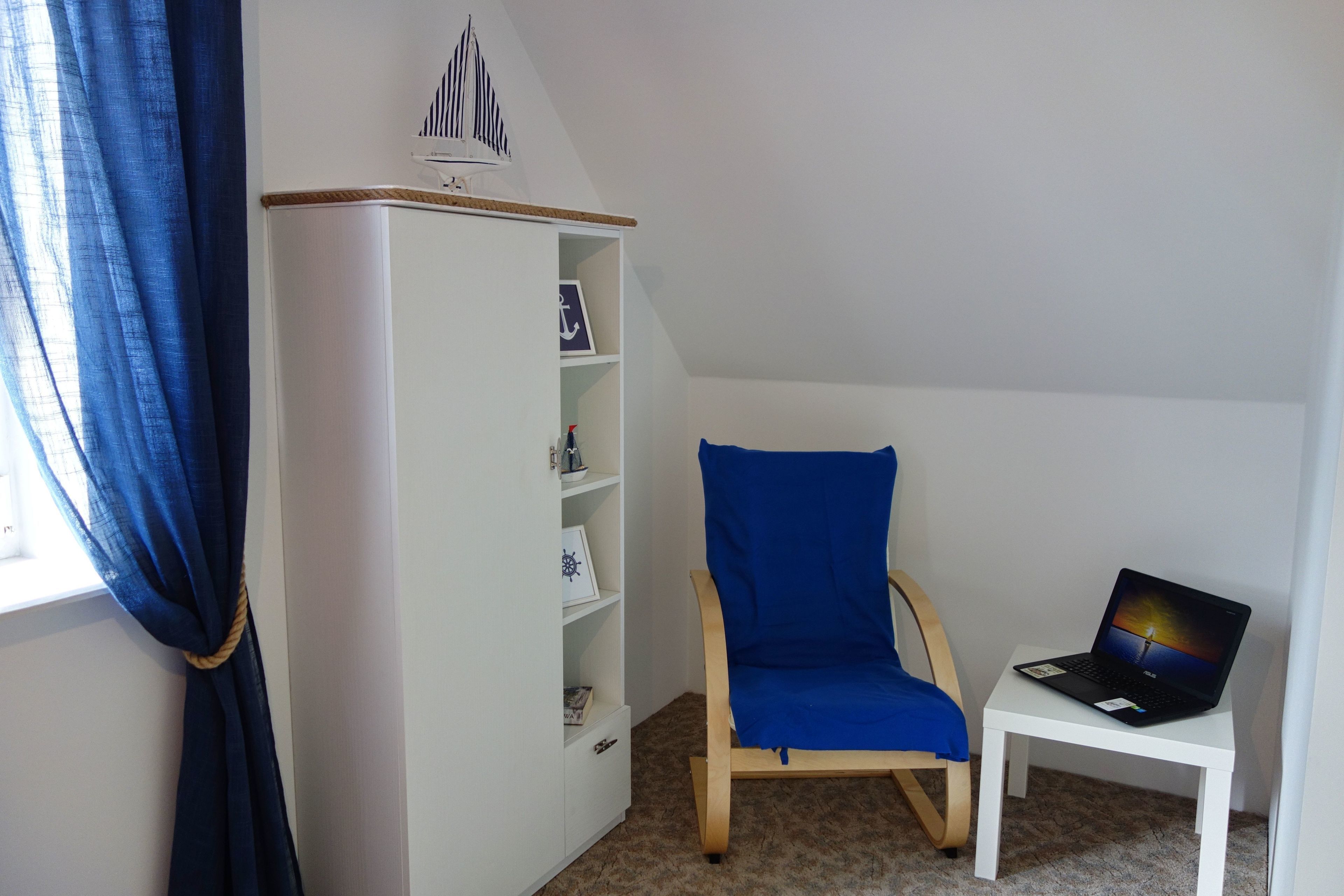 Une pièce avec une bibliothèque blanche, une chaise bleue et un ordinateur portable posé sur une petite table blanche.
