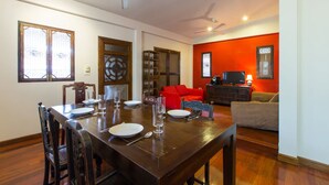 A dining area with a wooden table set for four, surrounded by chairs. There is a red sofa and a television in the living area. - Hua Hin Home (Hua Hin)
