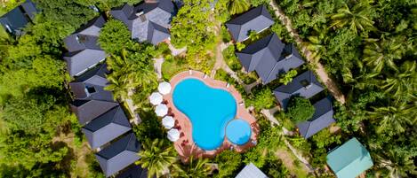 An aerial view of a resort with multiple bungalow-style buildings, a central swimming pool, and lush tropical vegetation.