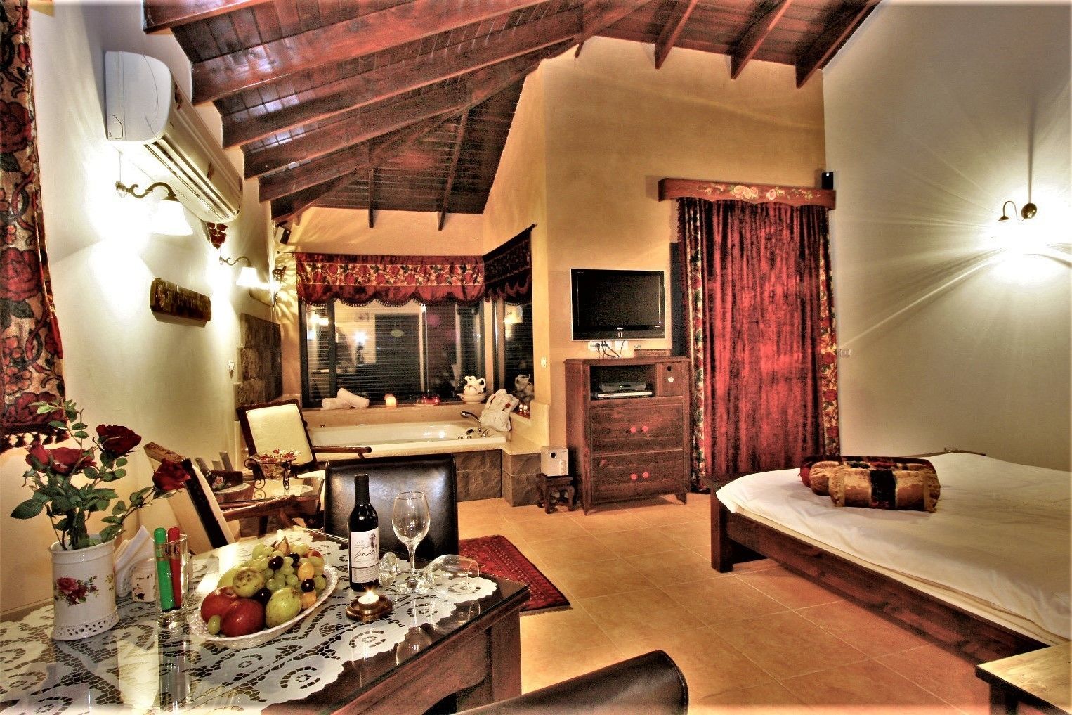 A cozy hotel room with a wooden ceiling, a bed, a TV, a small table with a fruit platter, and a window with curtains.