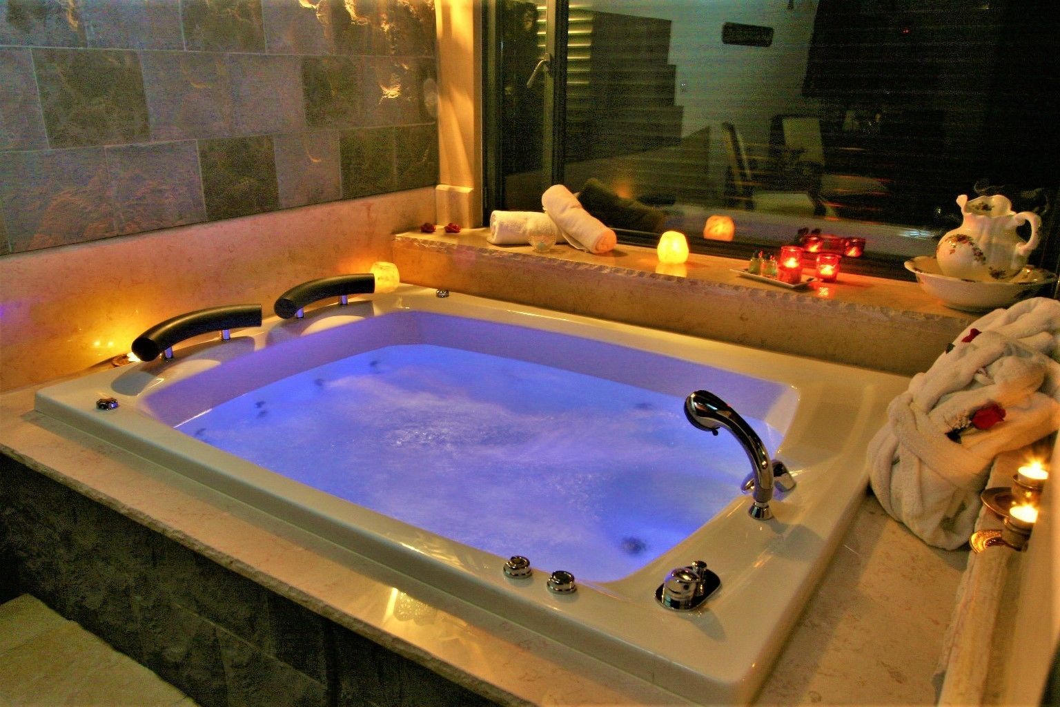 Een rechthoekig bubbelbad met een kraan en bedieningsknoppen, omringd door kaarsen en handdoeken op een aanrecht.