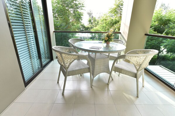 A balcony with a round glass table and wicker chairs, overlooking a lush green area.