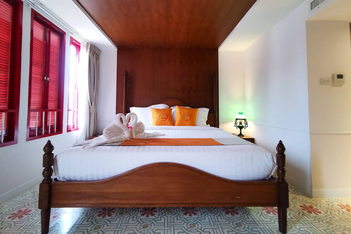Une chambre d’hôtel avec un lit en bois, des volets rouges, un sol à motifs et une table de chevet avec une lampe.