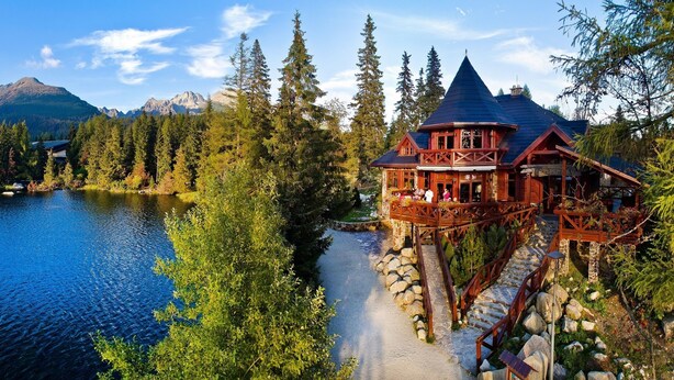 A lodge with a wooden exterior, a stone staircase, and a view of a lake and mountains. - Hotel Patria (Štrba)
