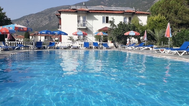 A swimming pool with lounge chairs and umbrellas, a building with a red roof, and mountains in the background. - Hotel Babadag (Fethiye)
