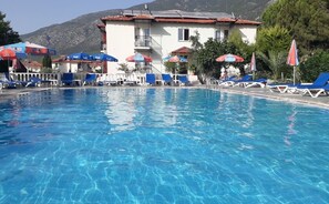 Una piscina con sillones de descanso y sombrillas, un edificio de techo rojo y montañas al fondo.