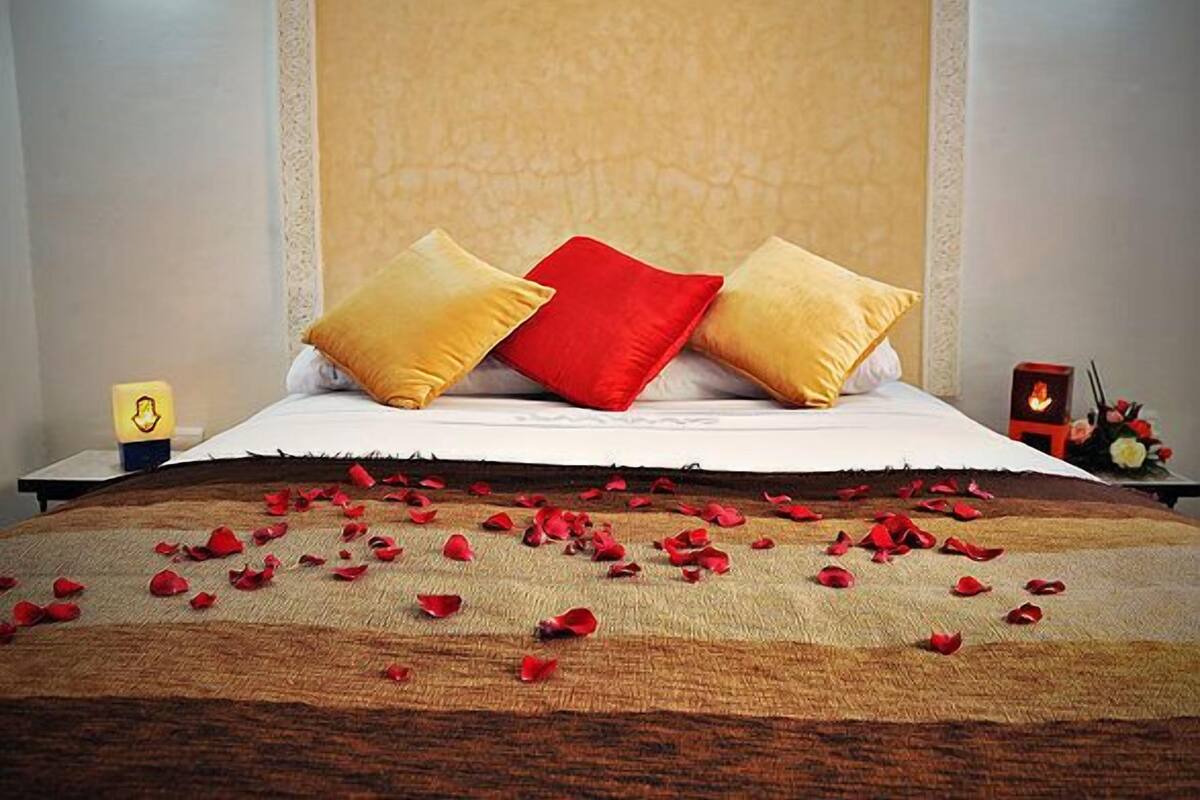 A neatly made bed with a headboard, a bedside table with a lamp, and scattered rose petals on the bed.