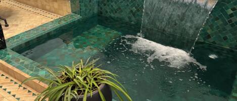 A pool with a waterfall feature, surrounded by a tiled area with a bench and cushions.