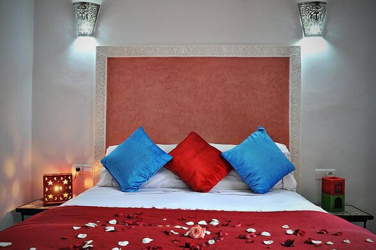 A bed with a red bedspread, two blue pillows, and a red pillow. There are rose petals on the bed.