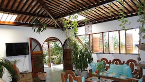 A covered patio area with wooden furniture, potted plants, and a television mounted on the wall. - Pousada Arco íris (Barra de Santo Antonio)