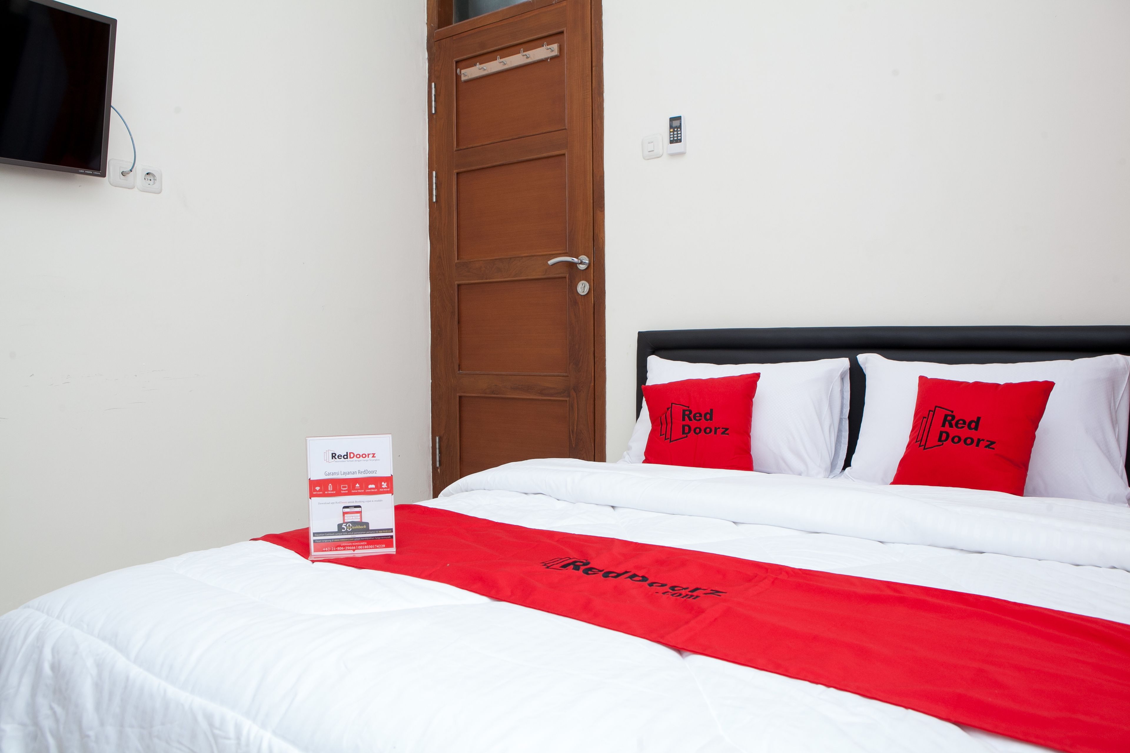 Una habitación de hotel con una cama, dos almohadas rojas, un edredón blanco y una colcha con la marca RedDoorz.