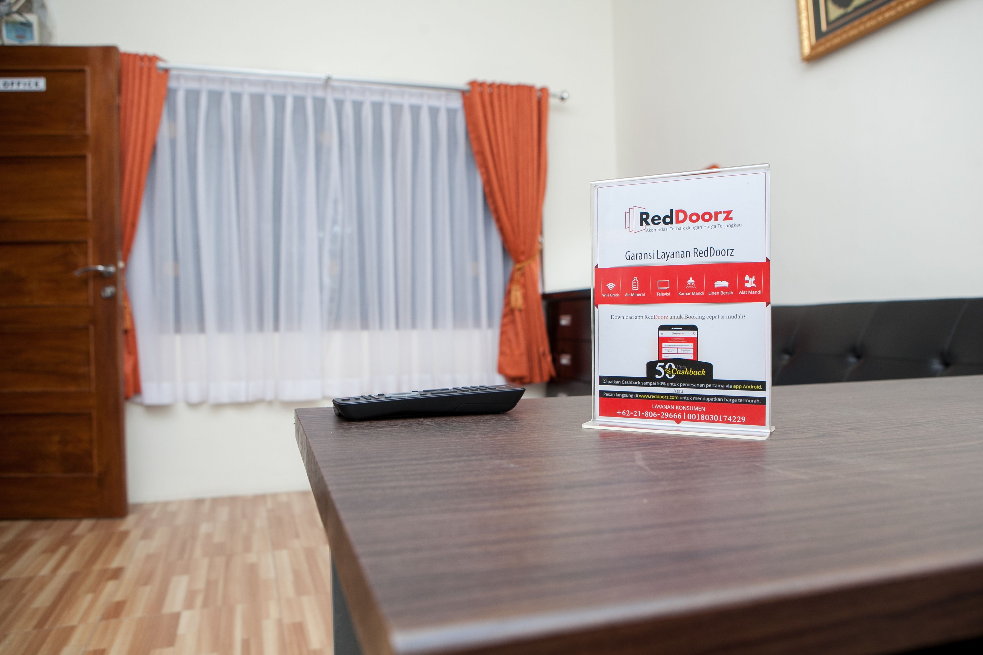 Ein RedDoorz-Schild auf einem Holztisch in einem Raum mit Fenster, Vorhängen und einer Fernbedienung.