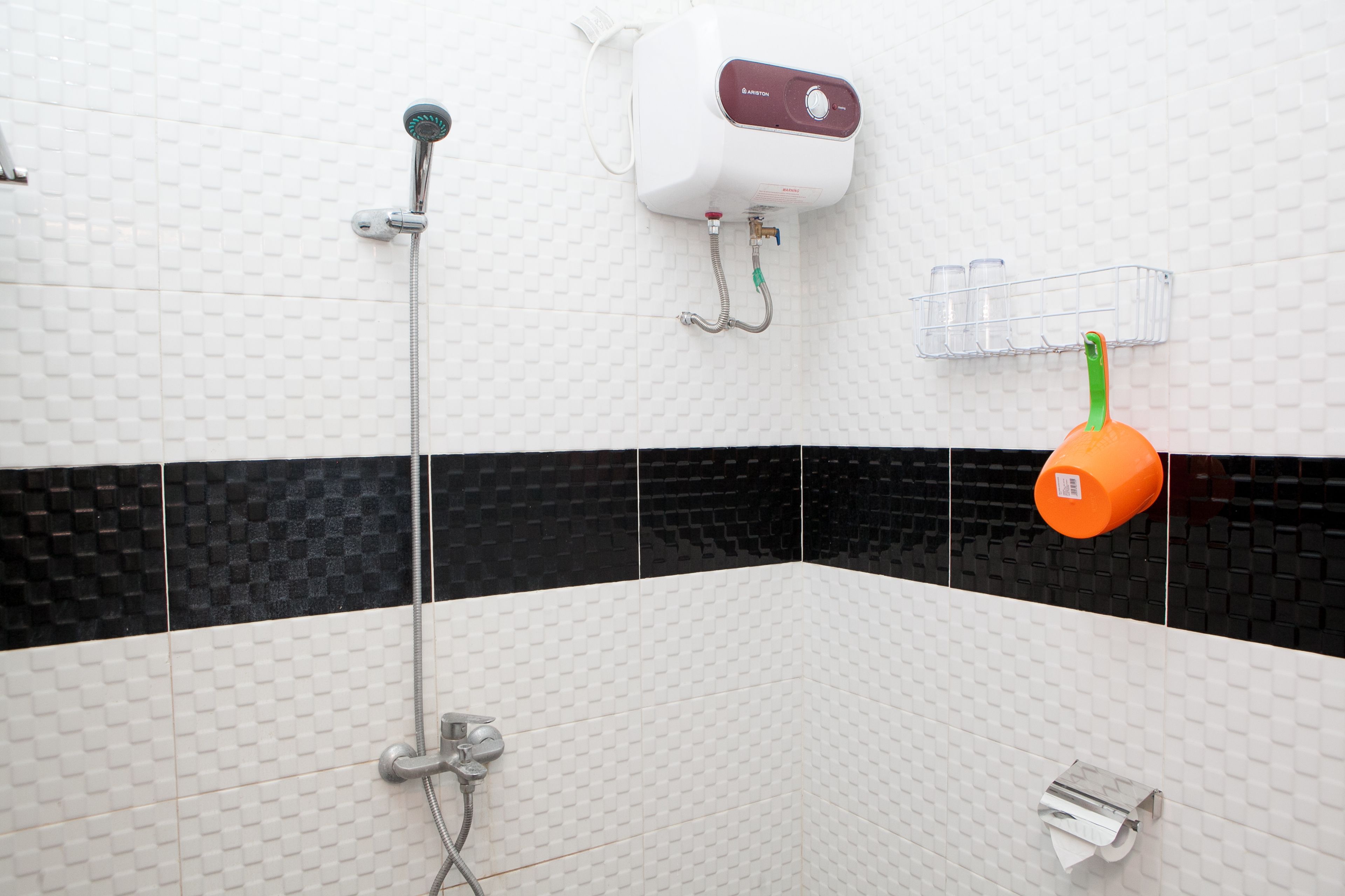 Un baño con pared de azulejos blancos y negros, un cabezal de ducha de mano, un calentador de agua instalado en la pared y un dispensador de jabón.