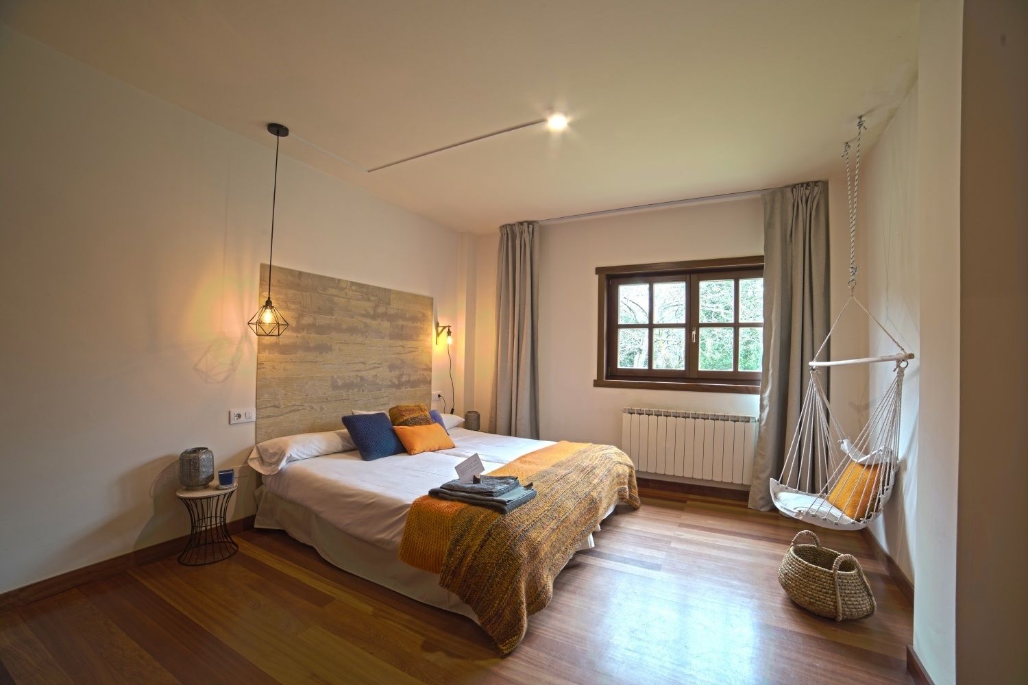 Un dormitorio con una cama grande, una silla colgante, una ventana con cortinas y suelo de madera.
