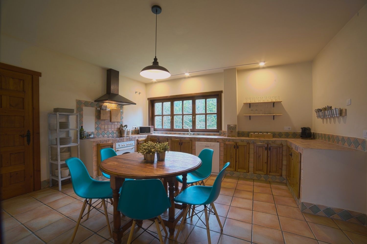 A kitchen with a round wooden table, four blue chairs, a hanging light fixture, a window, and a tiled backsplash.