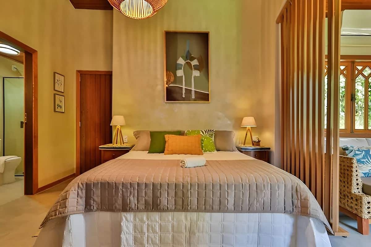 A bedroom with a large bed, two bedside tables with lamps, a painting on the wall, and a view through a window with a wooden lattice.