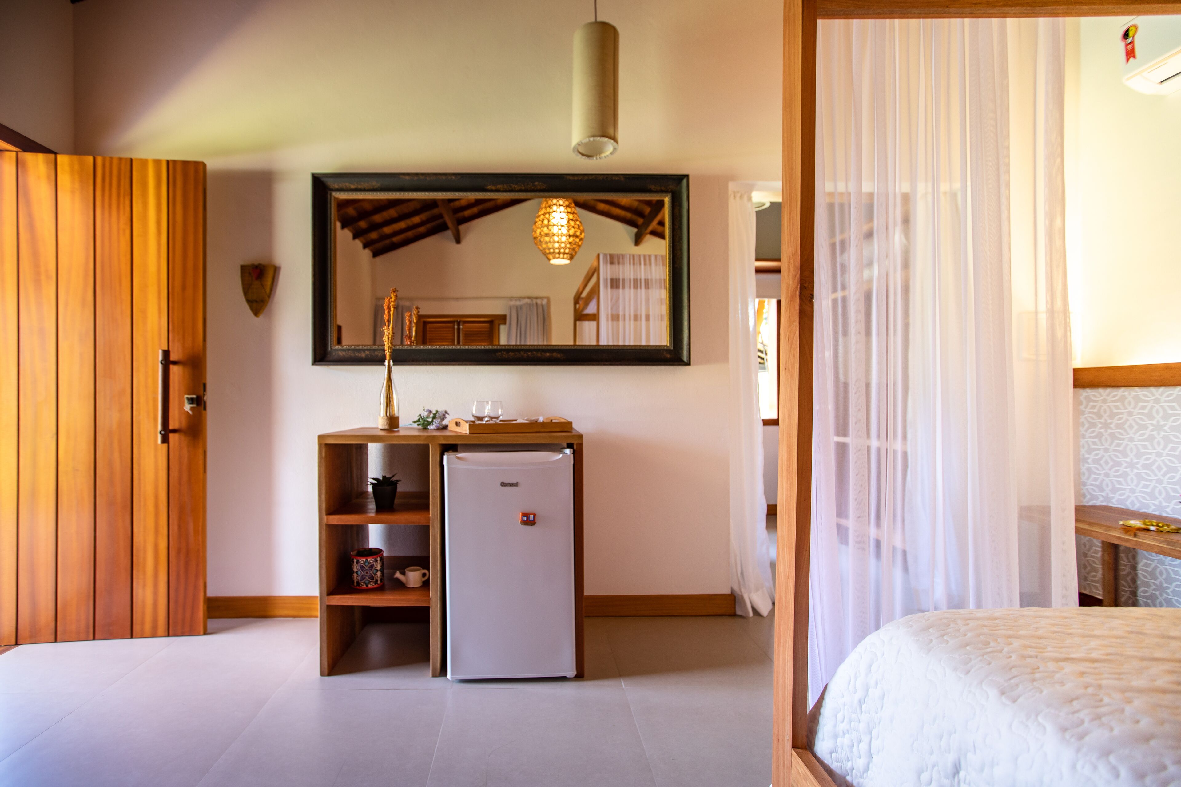 A bedroom with a bed, a wooden wardrobe, a small desk with a refrigerator, a mirror, and a hanging light fixture.