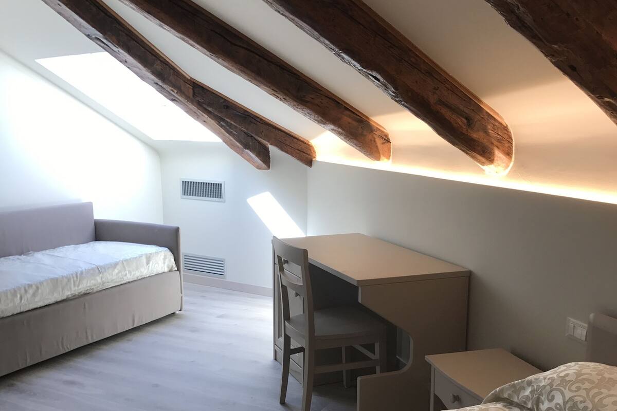 A bedroom with a loft bed, a desk, and a chair. There is a skylight above the bed and a vent on the wall.