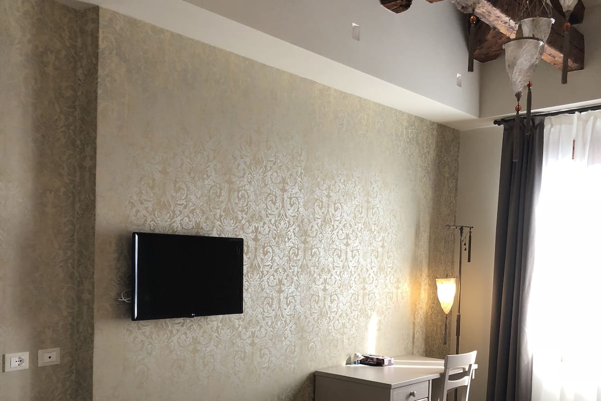 A hotel room with a bed, a desk, a chair, a TV mounted on the wall, and a window with curtains.