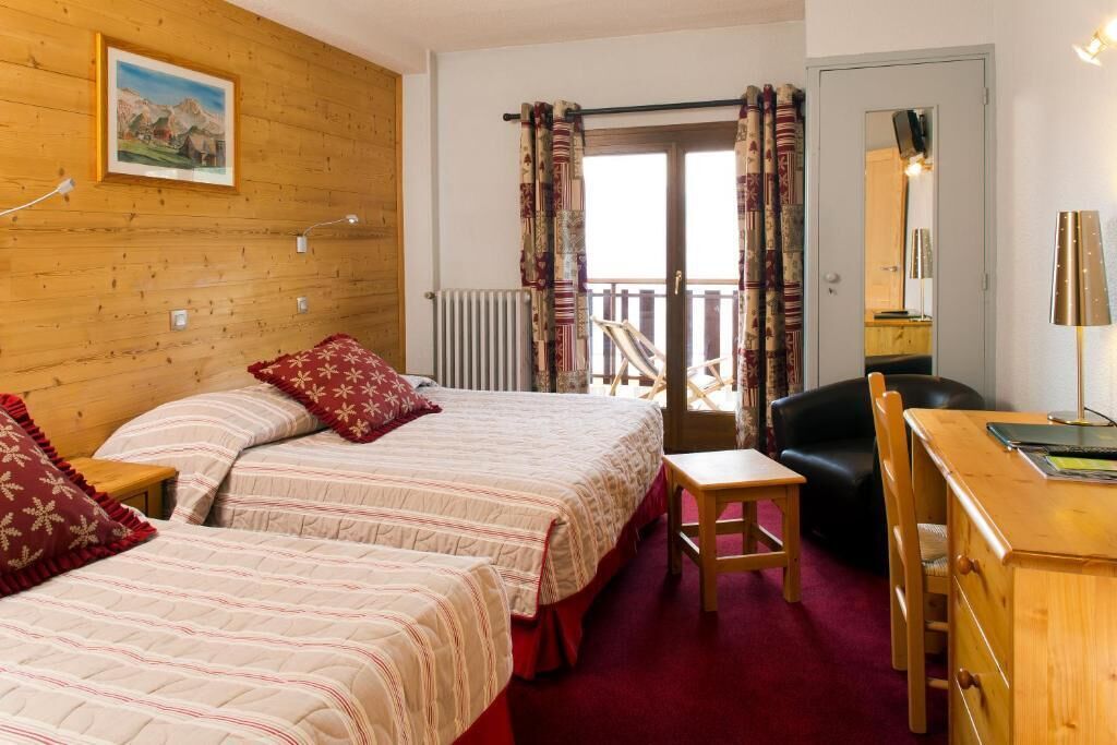 Een kamer met twee bedden, een bureau, een stoel, een radiator en een balkon met gordijnen.