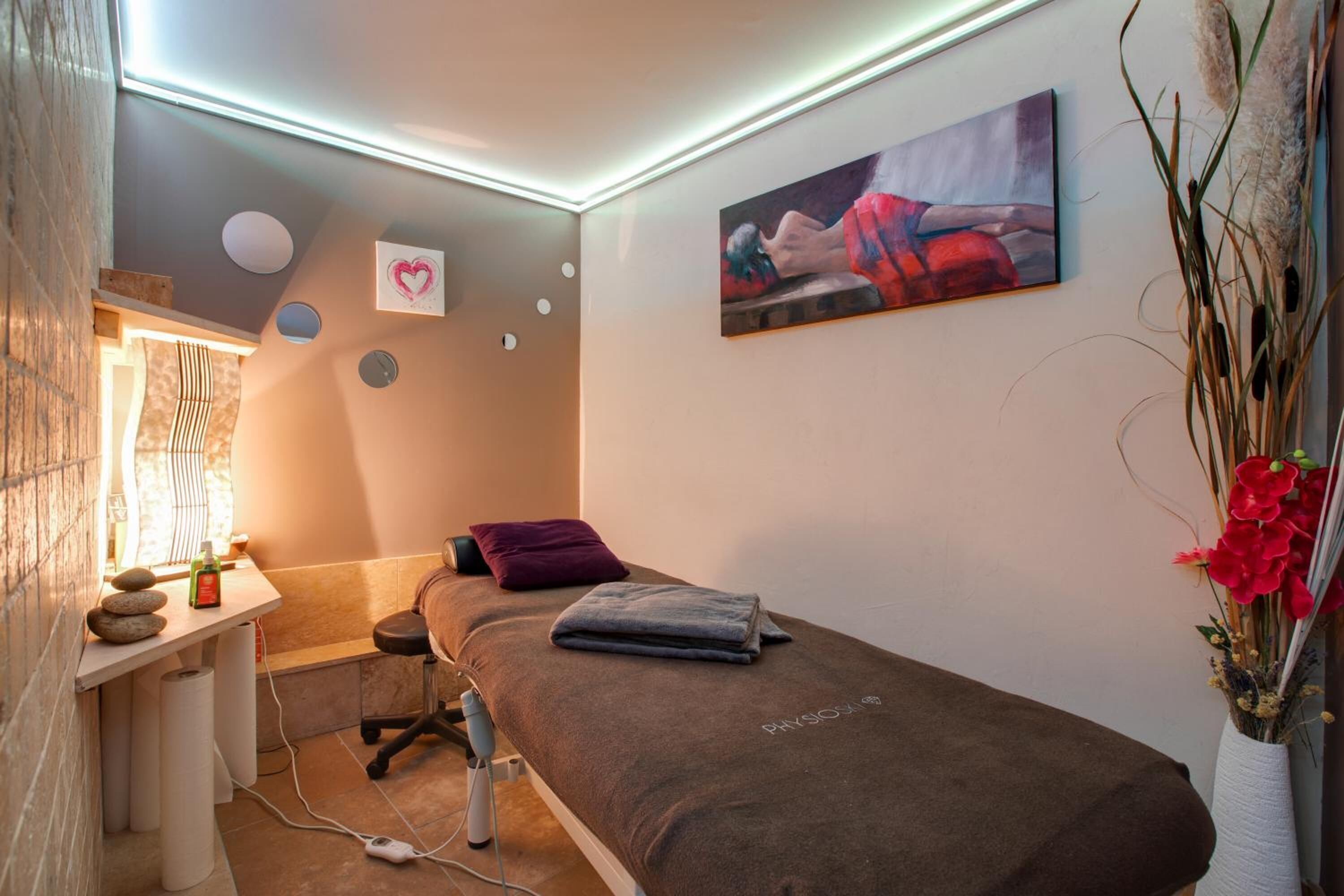 Ett rum för massagebehandlingar med en massagebänk, ett litet skrivbord med en flaska och några stenar, och en tavla på väggen.