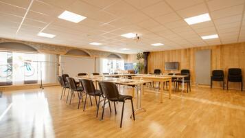 A spacious room with wooden flooring, multiple tables and chairs, and large windows allowing natural light.